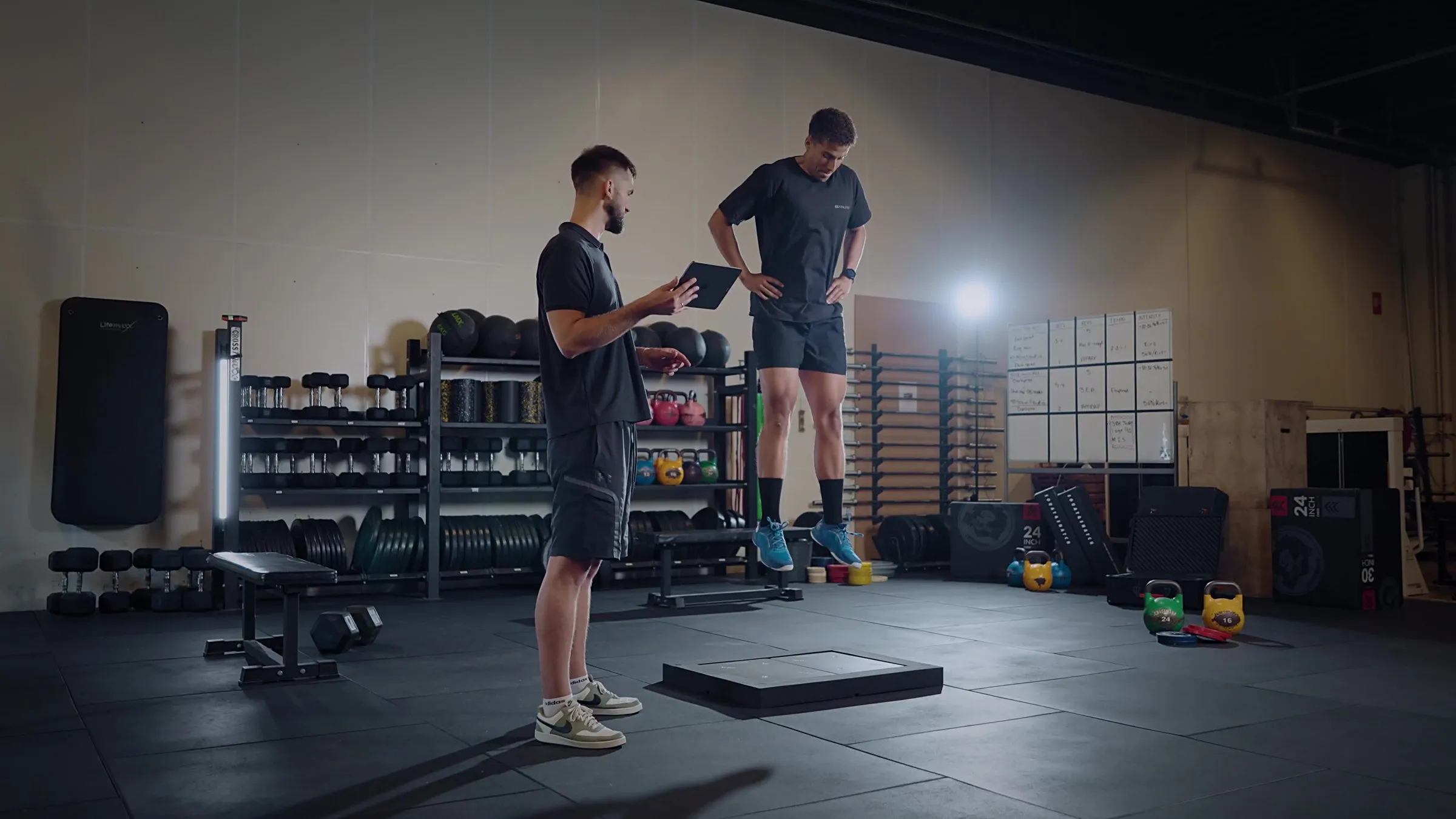 An athlete mid-air over a PlateMate force plate during a drop jump, watched by a coach holding a tablet.