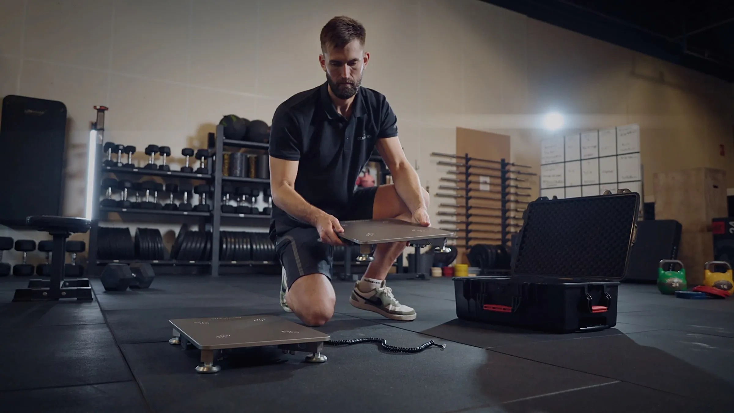 A coach kneeling on a gym floor, setting the second PlateMate force plate down next to the first, with the carry case open beside him.